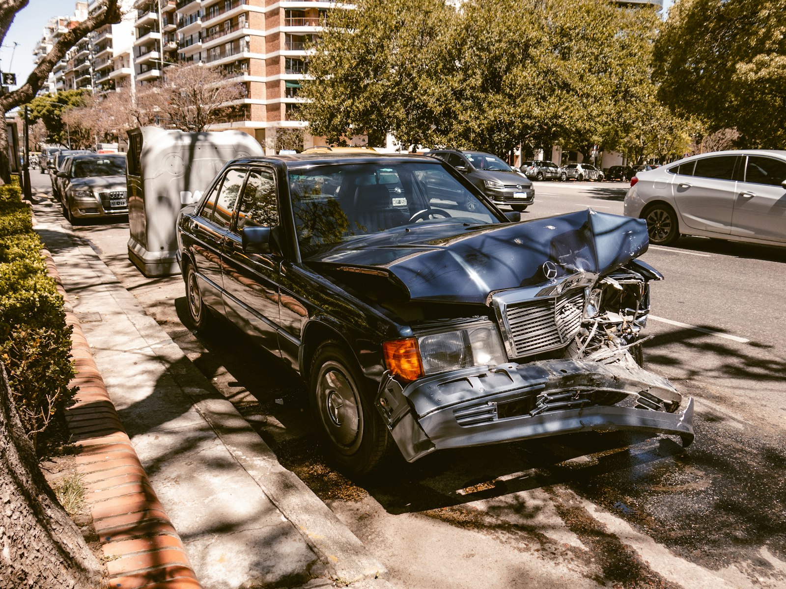Dark sedan with a crushed front end parked along a city street after a collision